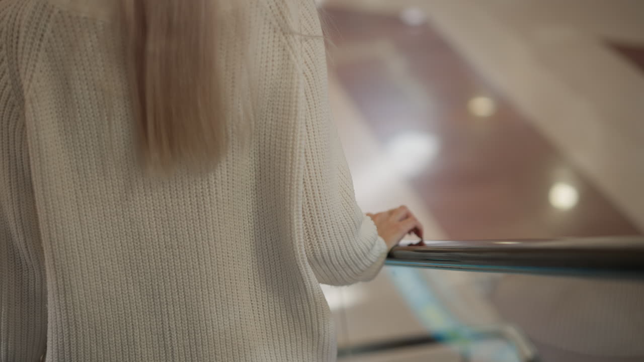 close up back view of stylish woman riding descending moving walkway in modern mall hall wearing white knit sweater and blonde hair flowing as blurred people move under bright store lights