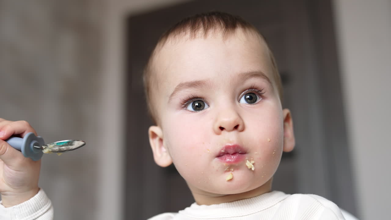 Grey-eyed Caucasian baby boy eating slowly from spoon. Some pieces of food falling from kid's cheeks. Low angle view close up portrait.