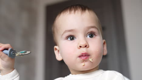 grey-eyed caucasian baby boy eating slowly from spoon. some pieces of food falling from kid's cheeks. low angle view close up portrait.