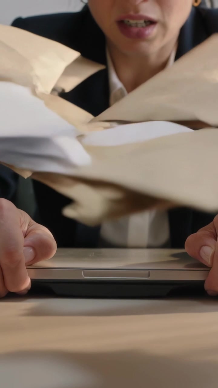 Vertical video: Bracing woman in blazer gripping laptop at desk, reacting to envelopes sweeping in