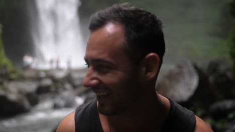 un joven en forma se ríe y sonríe hacia la cámara mientras está de pie con una hermosa y gran cascada en el fondo
