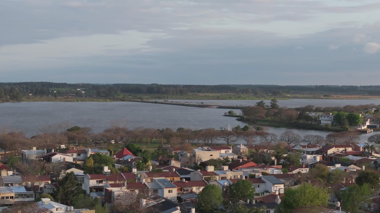 Aerial view of a tranquil riverside town with clustered houses amidst greenery under a vast cloudy sky, showcasing a blend of natural and urban landscapes in Federacion, Entre Ríos, Argentina