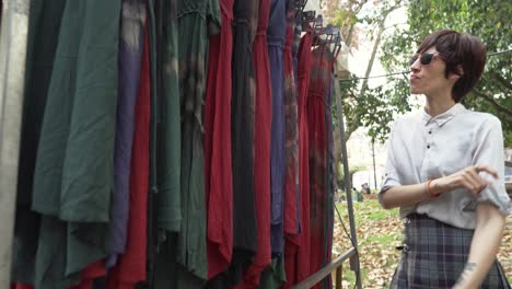 A-LGBTQ-woman-wearing-a-rainbow-bracelet-and-sunglasses-examines-different-dresses-at-a-street-market,-with-trees-in-the-background