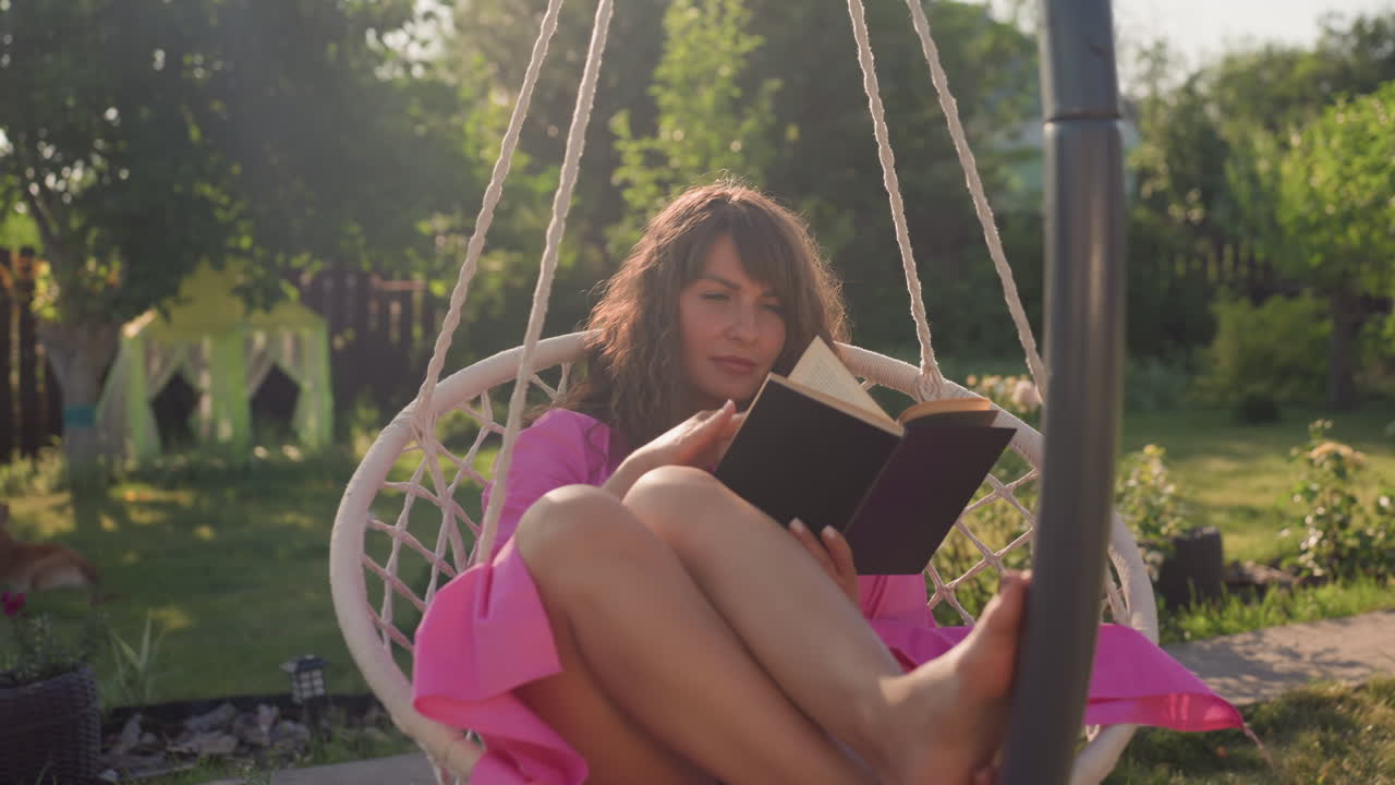 Peaceful Outdoor Reading, Tranquil Scene Of Woman Engrossed In Her Book Outdoors During Daylight, Calm Woman Peacefully Reading In Garden Swing Amid Gentle Afternoon Sunlight And Lush Greenery