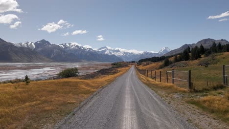 Empty-dirt-road-leading-towards-snowy-mountain-range-next-to-glacial-river-delta