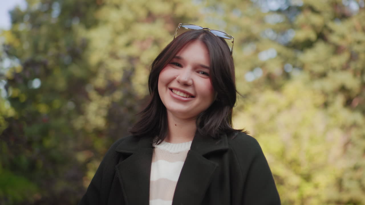 Retrato de una chica alegre con una sonrisa brillante, gafas de sol en el pelo, fondo suave bokeh de follaje, abrigo cómodo y suéter a rayas, luz natural, ambiente relajado al aire libre, expresión amigable, aire juvenil