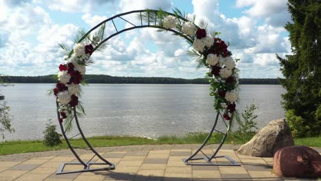 Vuele-A-Través-Del-Arco-De-Flores-Circular-De-La-Ceremonia-De-Boda-Hasta-Una-Vista-Al-Lago-En-Un-Día-Soleado