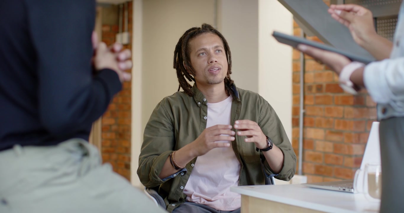 Afro-Amerikaans team presenteert tablet, zittende man luistert, gebaart, denkt achter bureau met laptop
