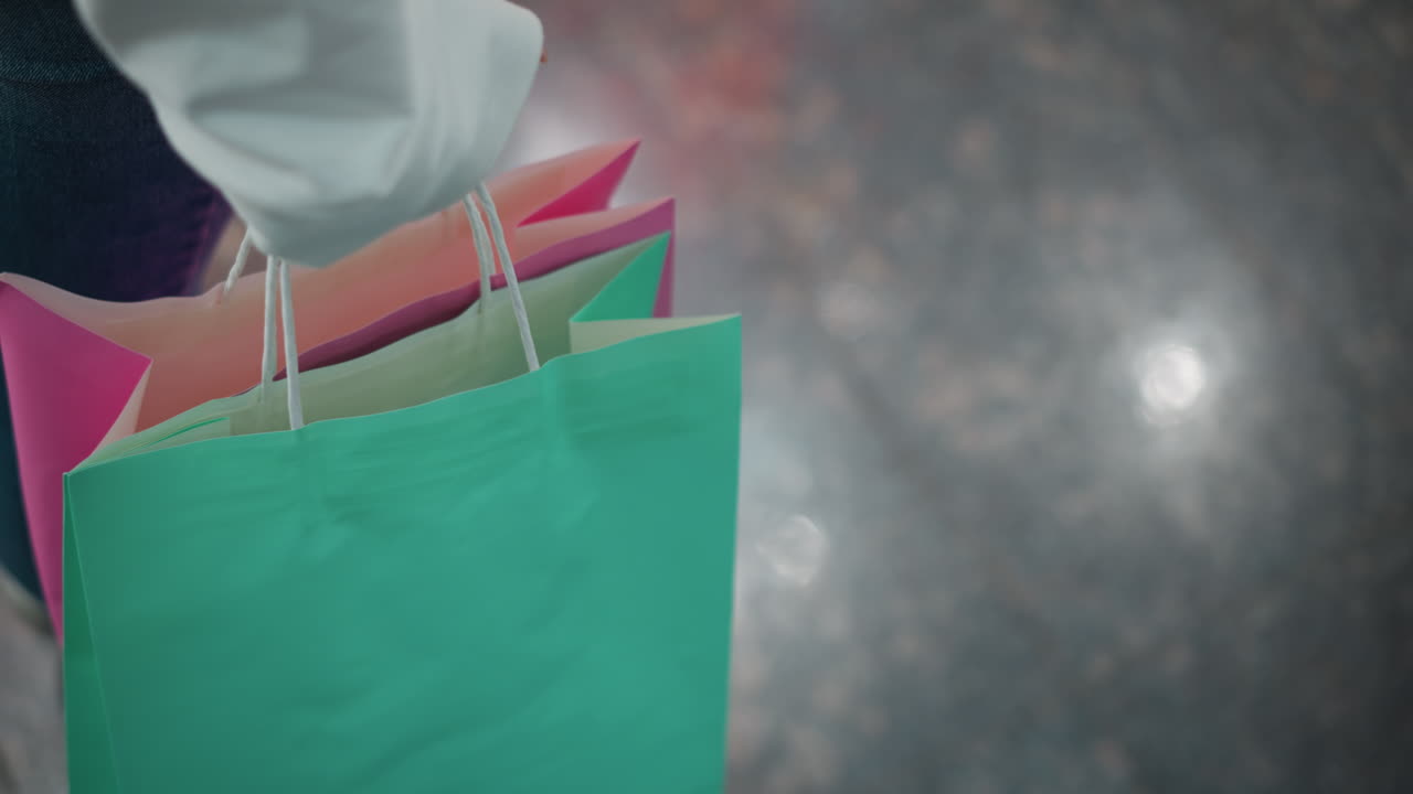 도시 환경에서 미묘한 움직임을 반영하는 밝은 조명 쇼핑몰 내의 반이는 대리석 바닥에서 는 동안 분홍색과 녹색 쇼핑 가방을 들고 있는 사람의 부분적 시각