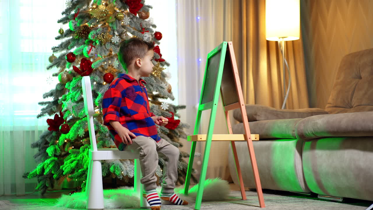 Lovely kid stands at the board near the Christmas tree. Baby boy draws with a chalk on the board