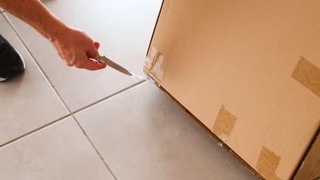 a person uses a knife to open a taped cardboard box on a tiled floor