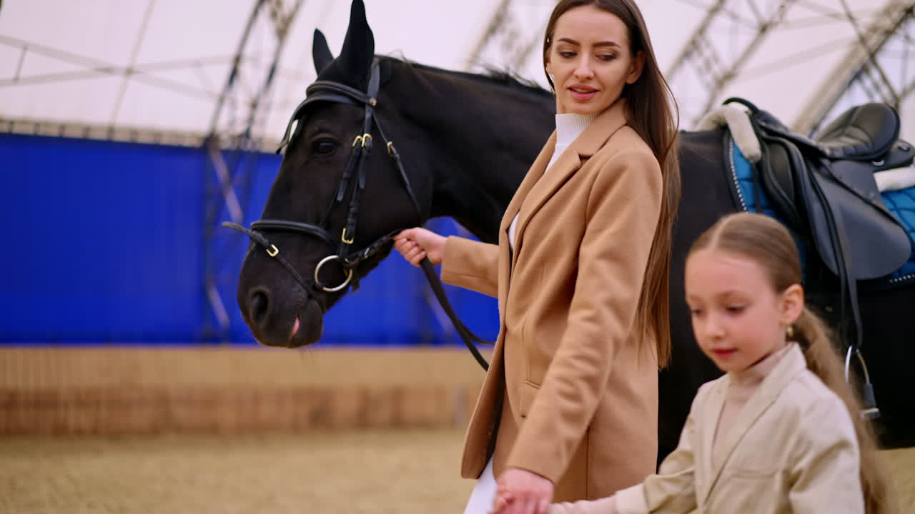 Attractive lady holding a girl by the hands leads a black horse in saddle and ammunition. Large riding hall at backdrop in blur.
