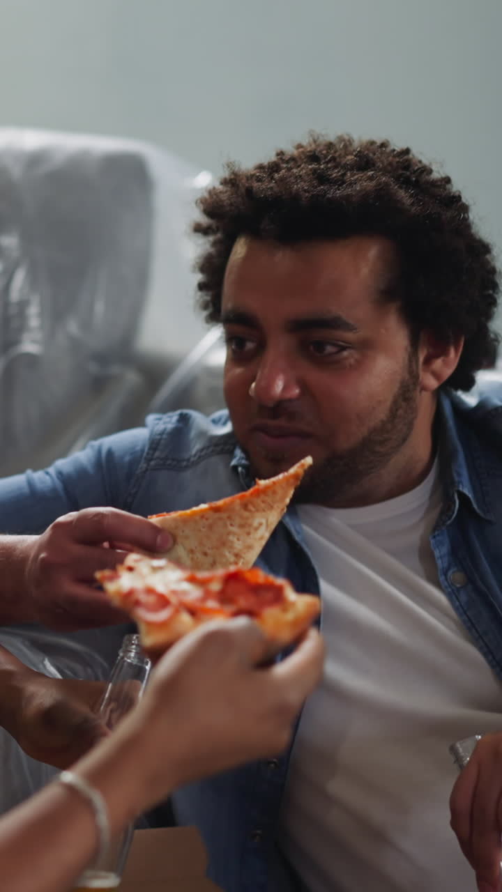 Cheerful African-American man eats pizza slice and talks to friend sitting near foiled sofa closeup. Young builders rest after repairing works in flat