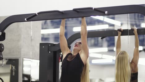 Athletic young blond female training on horizontal bar in black sportswear in gym performing pulls-up