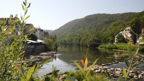 Blick-Auf-Den-Fluss-Nahe-In-Bad-Kreuznach-Mit-Einer-Hängebrücke-über-Dem-Wasser-Und-Menschen,-Die-Eine-Bootsfahrt-Genießen