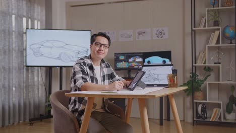 hombre asiático sonriendo y mostrando los pulgares hacia arriba gesto a la cámara mientras dibuja nuevo concepto de diseño de coche en una tableta en el estudio con televisión y computadoras exhibir modelo de coche eléctrico 3d