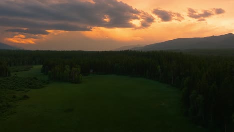 Drone-Aéreo-Volando-A-Través-De-Un-Exuberante-Campo-Verde-Rodeado-De-árboles-Altos-En-Las-Montañas-De-Montana-Durante-La-Impresionante-Puesta-De-Sol-Naranja-Y-Amarilla-Nublada