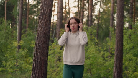 woman puts on headphones walking in forest. woman does sports running past trees and listening to favourite tracks jogging in wild on sunny summer day