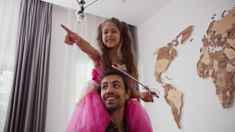 A-little-brunette-girl-in-a-fairy-costume-with-a-magic-wand-sits-on-the-shoulders-of-her-Happy-father-and-they-walk-around-their-modern-apartment