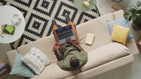 hombre afroamericano usando computadora portátil y bebiendo té en casa