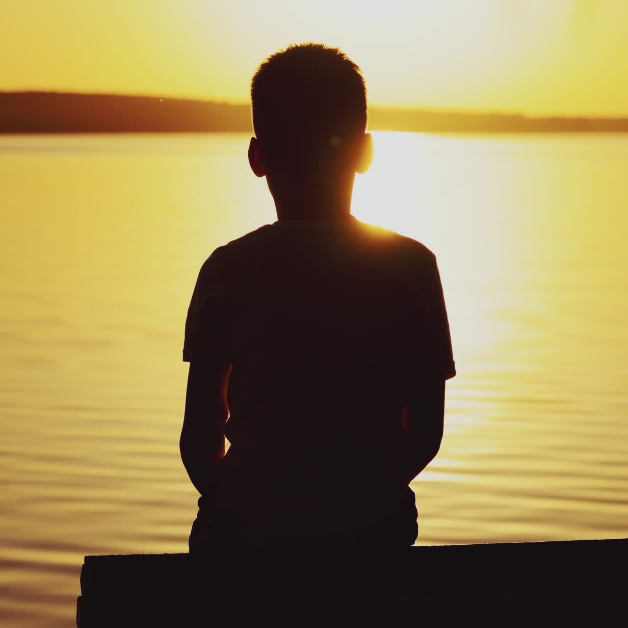 Little boy sitting on wooden dock at sunset. Child is watching the sunset against the background of a river. Dreams Come True.