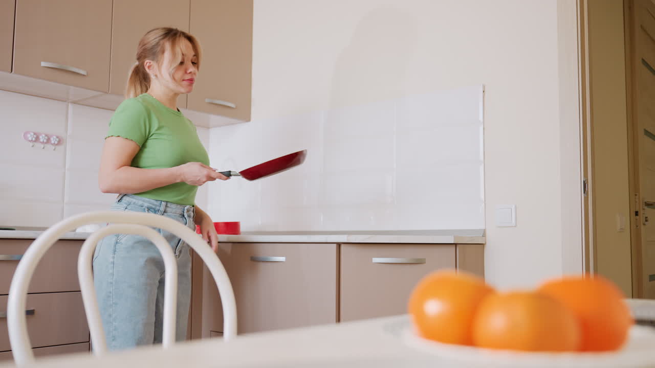 Chef in green shirt and jeans collects frying pan from open drawer in bright kitchen interior, with countertop visible and bowl of oranges in soft focus foreground creating homely lifestyle scene