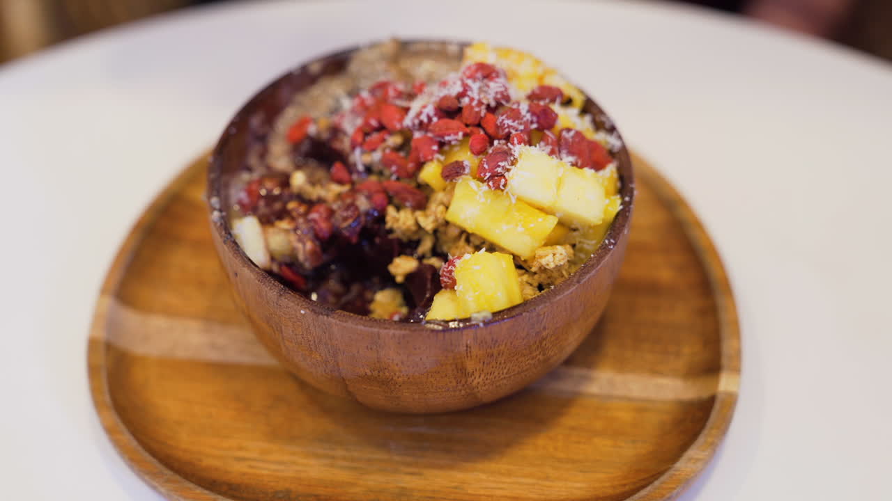 Close-up of wooden bowl with delicious acai bowl, topped with pineapple, mango, goji berries, granola and coconut flakes, being enjoyed by a customer.