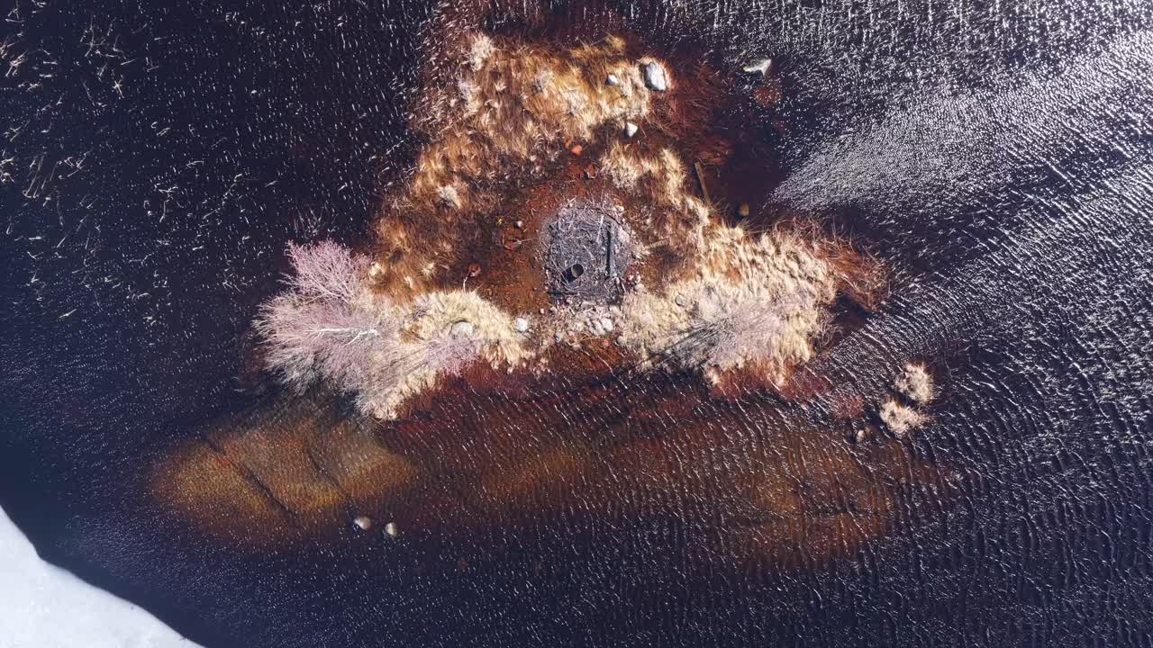 Rippled water around rocky shallow shoal. Lake partially covered in ice. Sunny Spring weather. Charred fire pit in center. Aerial top down view.