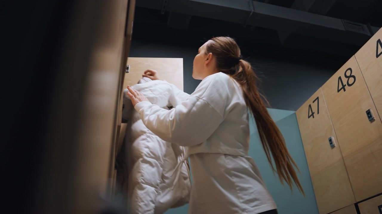 Locker badminton player zips puffer jacket before leaving, quick movements in numbered wooden locker area, focused expression, warm breath, light shadow play, brief celebration and relief after match