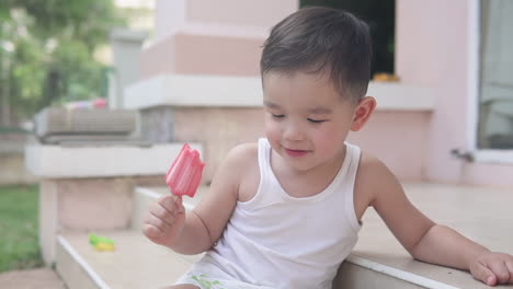 Video-En-Cámara-Lenta-De-Un-Niño-Pequeño-Asiático-Con-Ropa-De-Verano-Sentado-En-Las-Escaleras-Afuera-De-Una-Casa-Sosteniendo-Un-Helado-De-Paleta-De-Fresa-Roja-Sonriendo-Y-Pensando