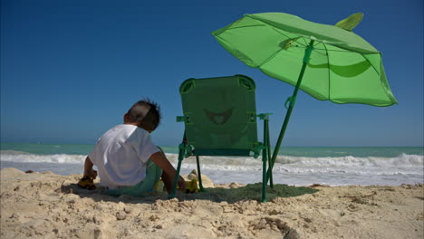 movimiento lento de un joven niño latino jugando solo en la arena junto a su silla de playa y paraguas en un hermoso día soleado