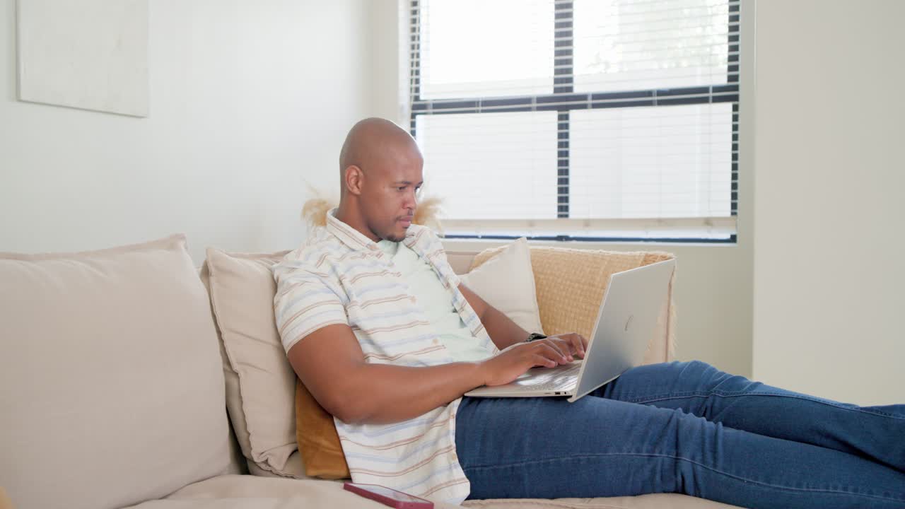 African American man at home on sofa, typing on laptop, pausing, checking smartphone while working