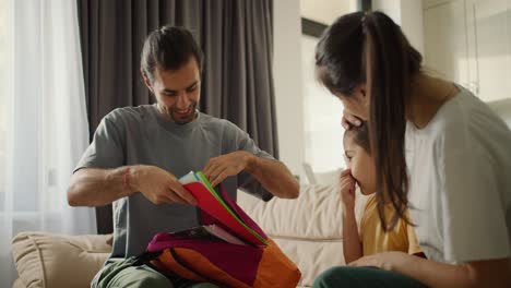 Happy-young-parents-are-getting-their-daughter-ready-for-school-and-packing-a-backpack-for-her,-they-put-different-notebooks-and-textbooks-there-on-a-light-brown-sofa-in-a-modern-room