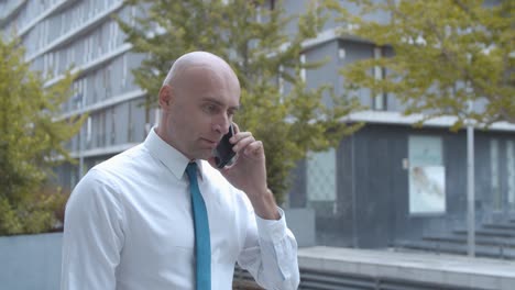 dolly shot of a confident businessman in white shirt, talking on mobile phone while walking near office building outside