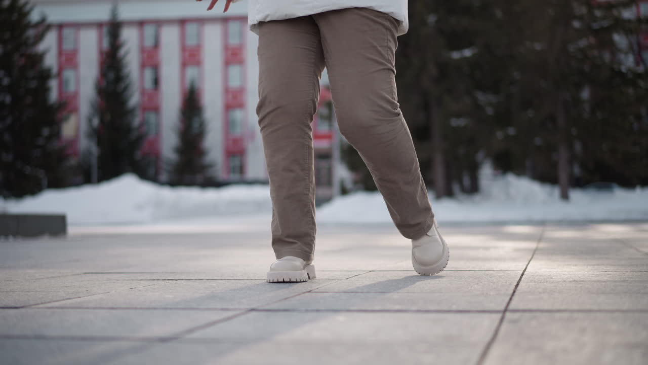 Leg view of tap dancer wearing brown trousers and canvas boots tapping rhythmically on large outdoor pavement tiles under winter light casting long shadows creating dynamic movement energy