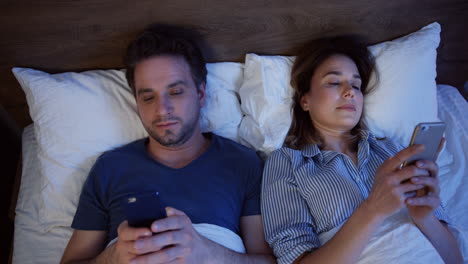 Top-view-of-caucasian-couple-lying-on-the-bed-and-using-smartphones-at-night