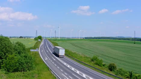 Großer-Weißer-Transportwagen,-Der-Auf-Einer-Landstraße-Mit-Einem-Riesigen-ökologischen-Windpark-Im-Hintergrund-Fährt