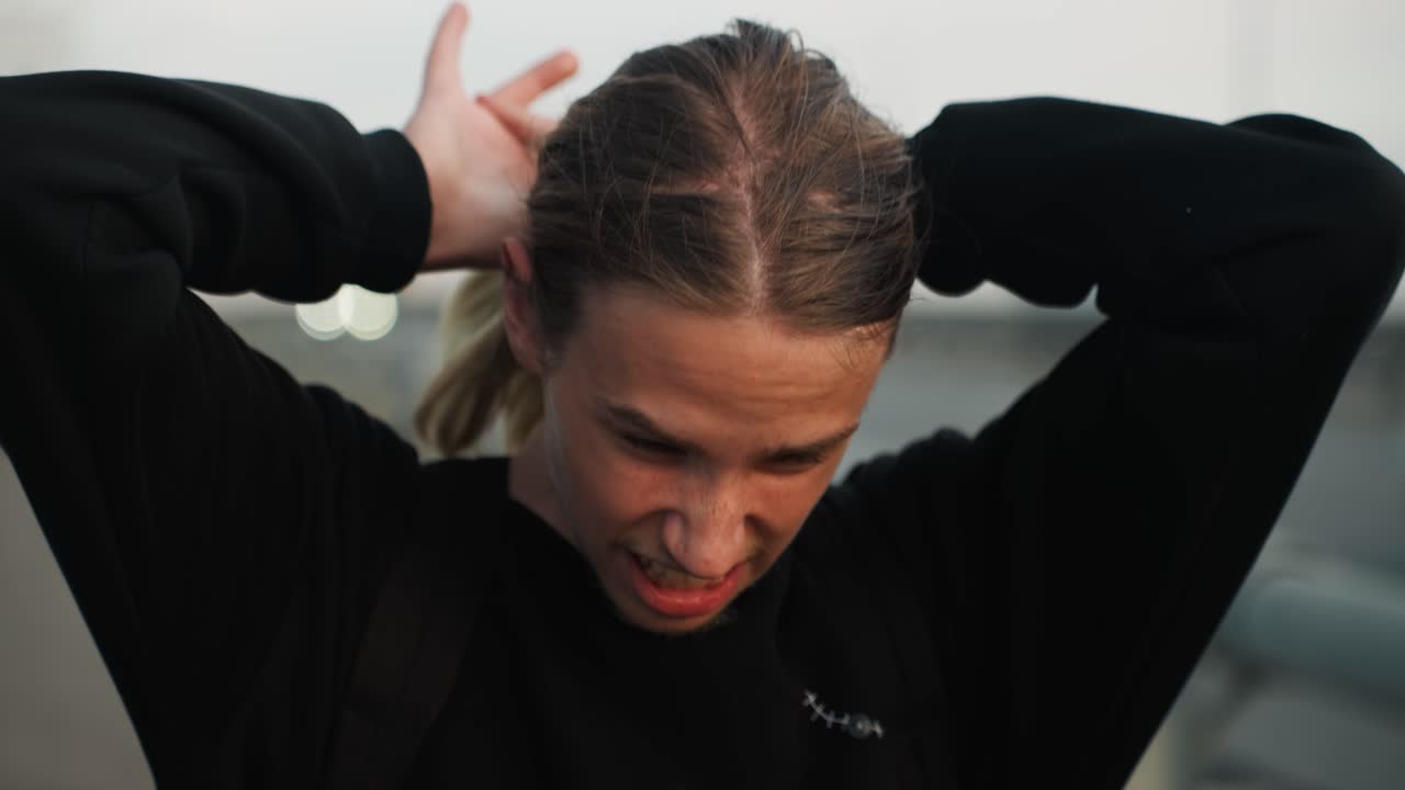 Focused young man braiding hair outdoors, twilight overpass and highway in background, hands methodically tying cornrows into ponytail, backpack visible, concentrated expression and candid urban