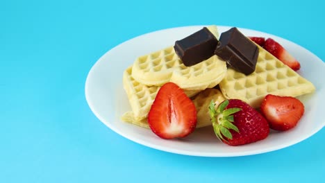 waffles on plate with two chocolate cubes and strawberries on blue background