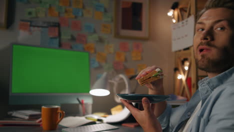 hungry student eating sandwich evening room. man taking break in front computer