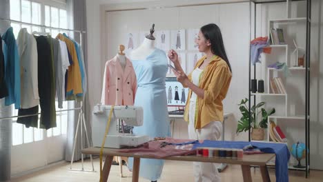 diseñadora asiática sonriente con máquina de coser mirando un vestido en un maniquí y comparándolo con las imágenes en el teléfono inteligente mientras diseña ropa en el estudio