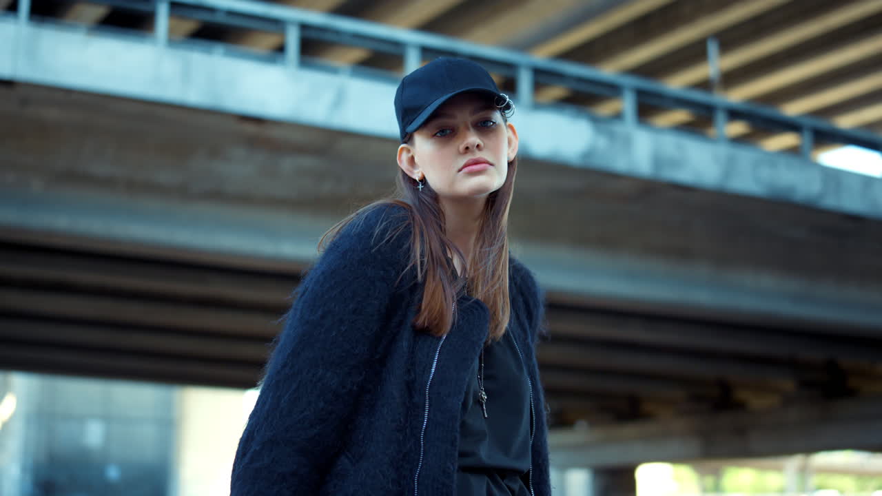 Attractive woman in cap standing on city street. Serious lady posing at camera