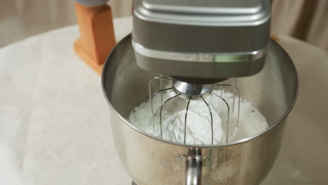 electric mixer with stainless whisk attachment filled with thick white dough resting inside metal bowl on kitchen table, additional kitchen tools visible in background