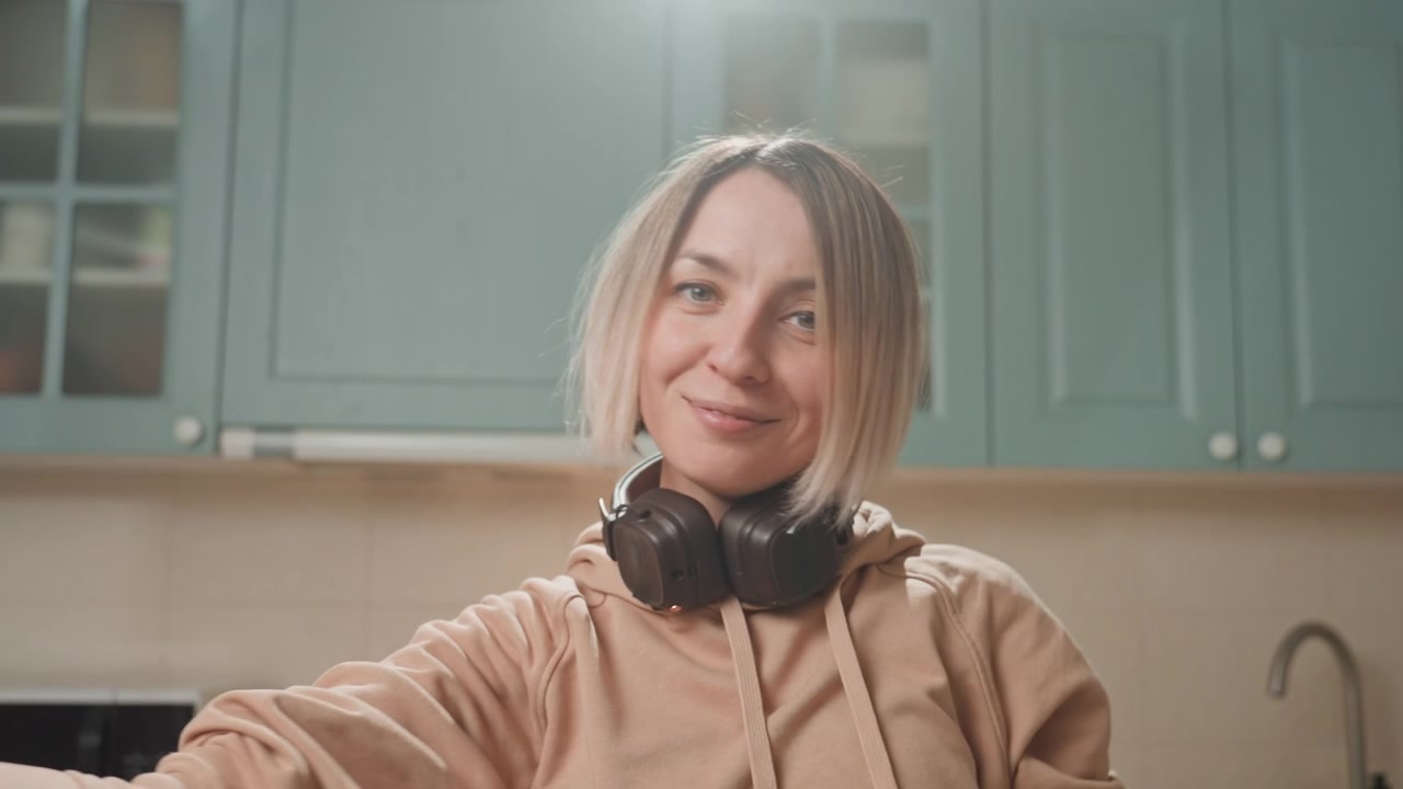 Blanke vrouw heft armen met koptelefoon in de keuken, extatische vreugdevolle ontlading met vloeiende gebaren en stralende glimlach, casual hoodie, warme tegenlicht, spontane thuisvoorstelling vol gelukzaligheid.