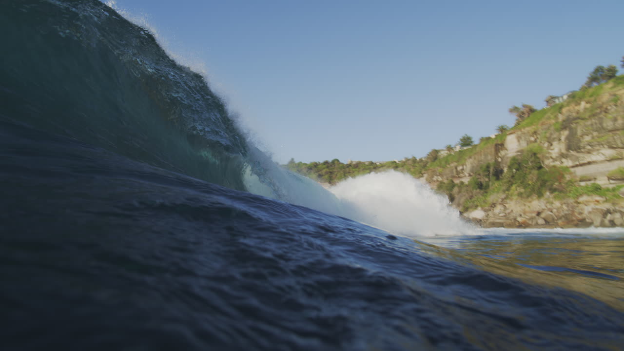 Slow-motion shot of a morning wave crashing against the shoreline, filmed from outside the water, highlighting the wave's force and spray