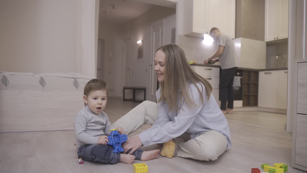 bebé jugando sentado en el suelo con juguetes y con su madre. al fondo su padre está en la cocina