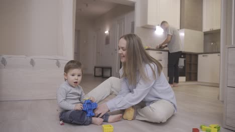 baby playing sitting on the floor with toys and with his mother. in the background his father is in the kitchen