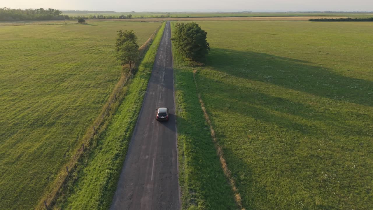 A solitary car travels along a straight, empty road cutting through expansive green fields. The aerial view captures the peaceful countryside and the sense of freedom in open space