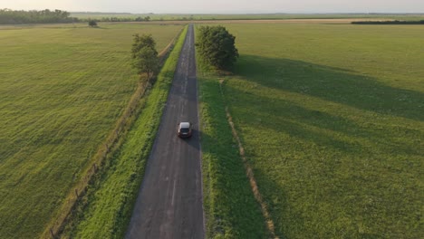 a solitary car travels along a straight, empty road cutting through expansive green fields. the aerial view captures the peaceful countryside and the sense of freedom in open space