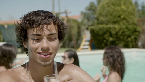 front view of man with naked torso standing in swimming pool, drinking while his friends dance in the pool